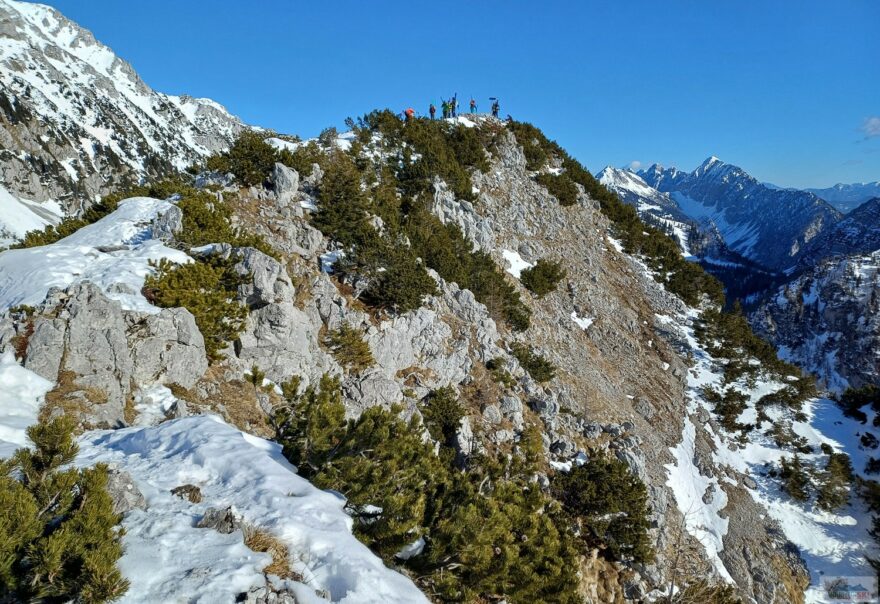 Skialpinisti na vrcholu Srednji vrh při pohledu od výhlídkové lavičky